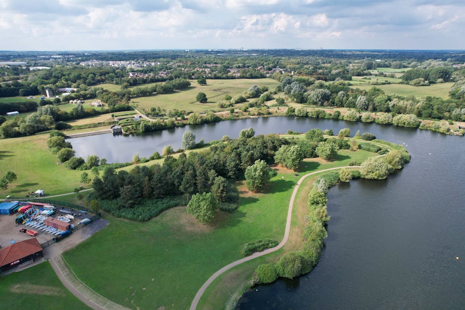An aerial view of a small lake surrounded by trees and greenery, with a narrow concrete pathway running along the right side, adjacent to a winding road. On the left side of the lake, there is a small