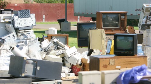 A large outdoor collection of discarded electronic waste, including old CRT televisions with wooden and plastic casings, computer monitors, and various small electronic devices, all piled on a paved surface during daytime. Some items are stacked haphazardly, with visible cables and circuit boards exposed. The background features a red brick wall, a strip of green grass, and a grey metal container or building, indicating a waste collection or clearance area in Hillingdon. The scene is illuminated by natural daylight, highlighting the variety of textures and colours of the electronic components and housings. This image reflects the type of bulky rubbish that Commercial Waste Removal Hillingdon manages, particularly focusing on electronic waste removal services in the local area, emphasizing the importance of proper disposal of obsolete gadgets and appliances.