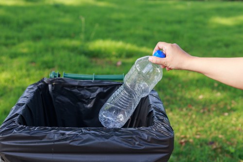 A person's hand is visible, holding a clear plastic water bottle with a blue cap, which is being positioned above a black plastic rubbish bin. The bin is located outdoors on a paved surface, with a well-maintained green grassy area in the background. The scene appears to be in daylight, with natural lighting highlighting the texture of the plastic bottle and the smooth, shiny surface of the bin. The plastic bottle is partially filled with water and is held upright, ready to be disposed of or recycled. The environment suggests a typical waste collection or rubbish disposal activity, possibly near a residential or commercial property in Hillingdon, aligning with the services offered by Commercial Waste Removal Hillingdon for rubbish collection and waste management.